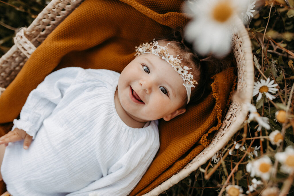 Auf dem Bild sieht man ein Baby. Es liegt auf einer braunen Decke in einem Korb mitten in einem Blumenfeld