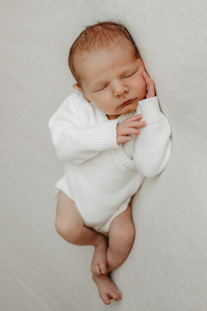 Kontakt. Familienfotografin Calw – Alyssa Rathfelder. Auf dem Bild sieht man ein Baby. Es trägt einen weißen Body und schläft auf einer beigen Decke