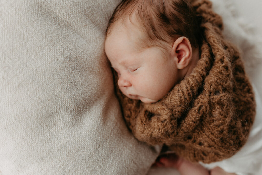 Auf dem Bild sieht man ein Baby, wie es auf einem beigen Kissen liegt. Es trägt einem braunen Pullover
