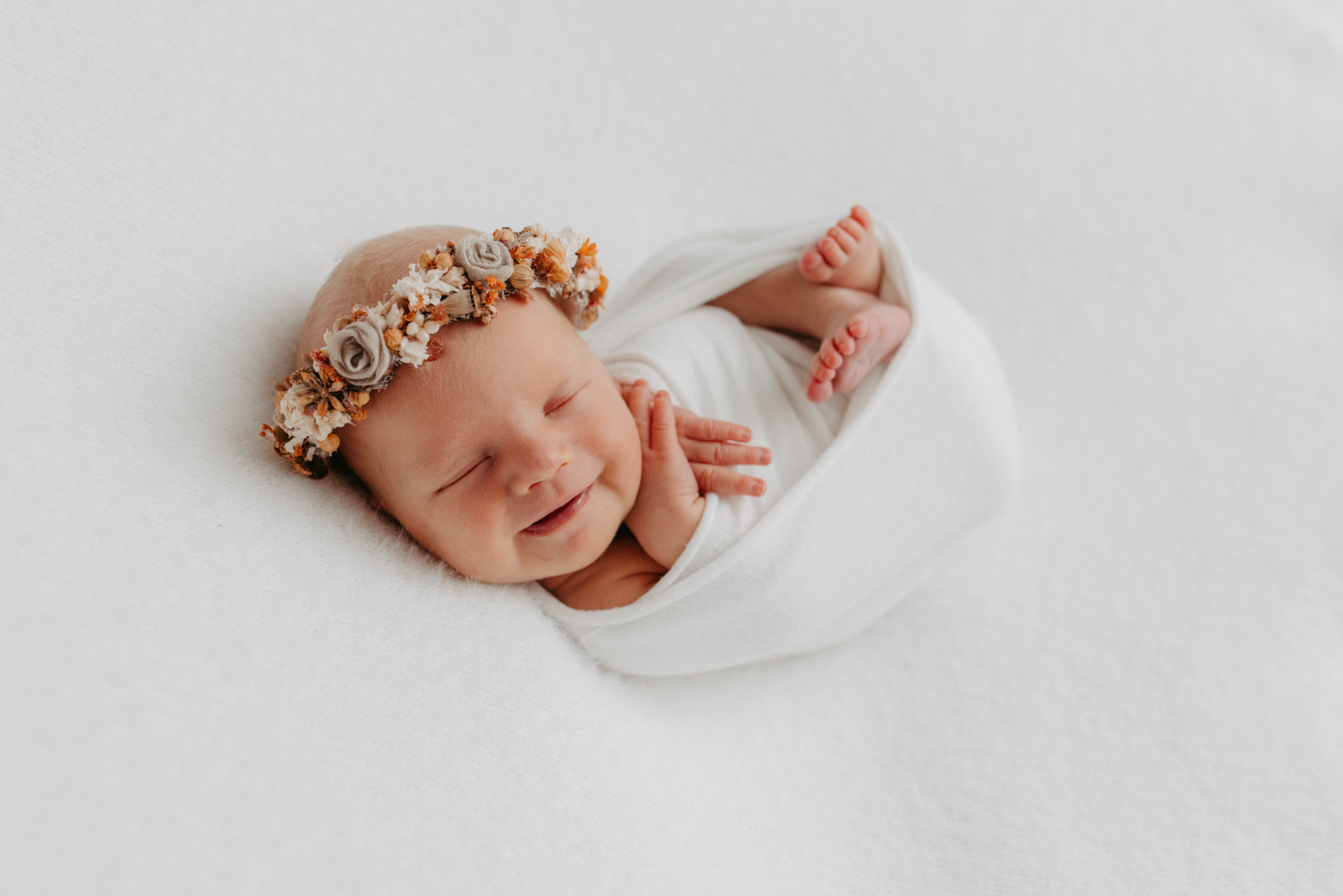 Auf dem Bild sieht man ein lächelndes Baby. eingepuckt in ein weißes Tuch. Es trägt einen Blumenhaarkranz