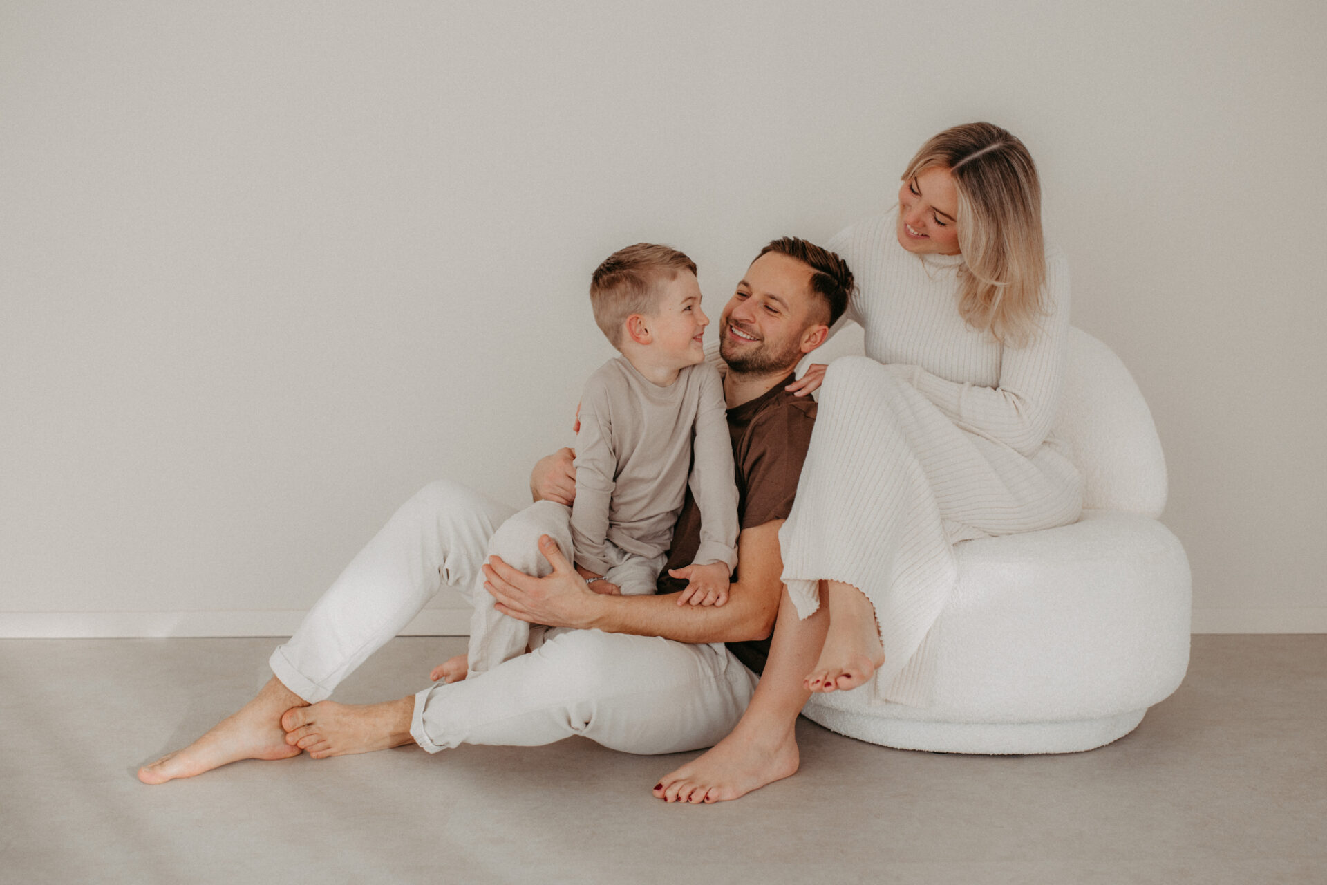 Familienshooting Calw. Auf dem Bild sieht man eine dreiköpfige Familie. Sie schauen sich gegenseitig an. Das Bild ist in einem Studio. Sie tragen alle helle Farben und sitzen auf einem beigen Stuhl